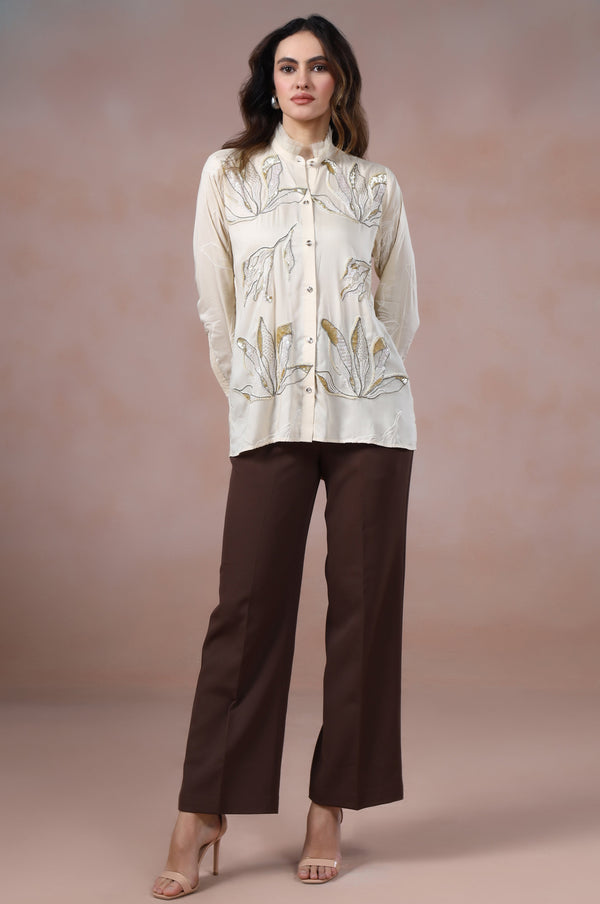 Woman wearing a cream blouse with floral patterns and brown pants against a pink background