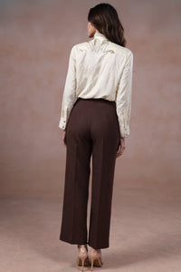 Woman wearing a white blouse and brown pants against a beige background