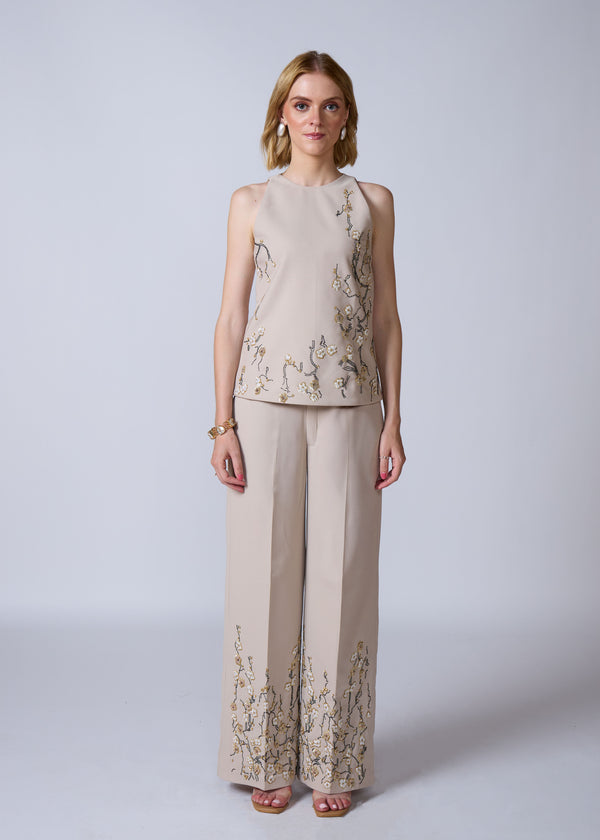 Woman wearing a beige sleeveless top and wide-leg pants with floral embroidery on a white background
