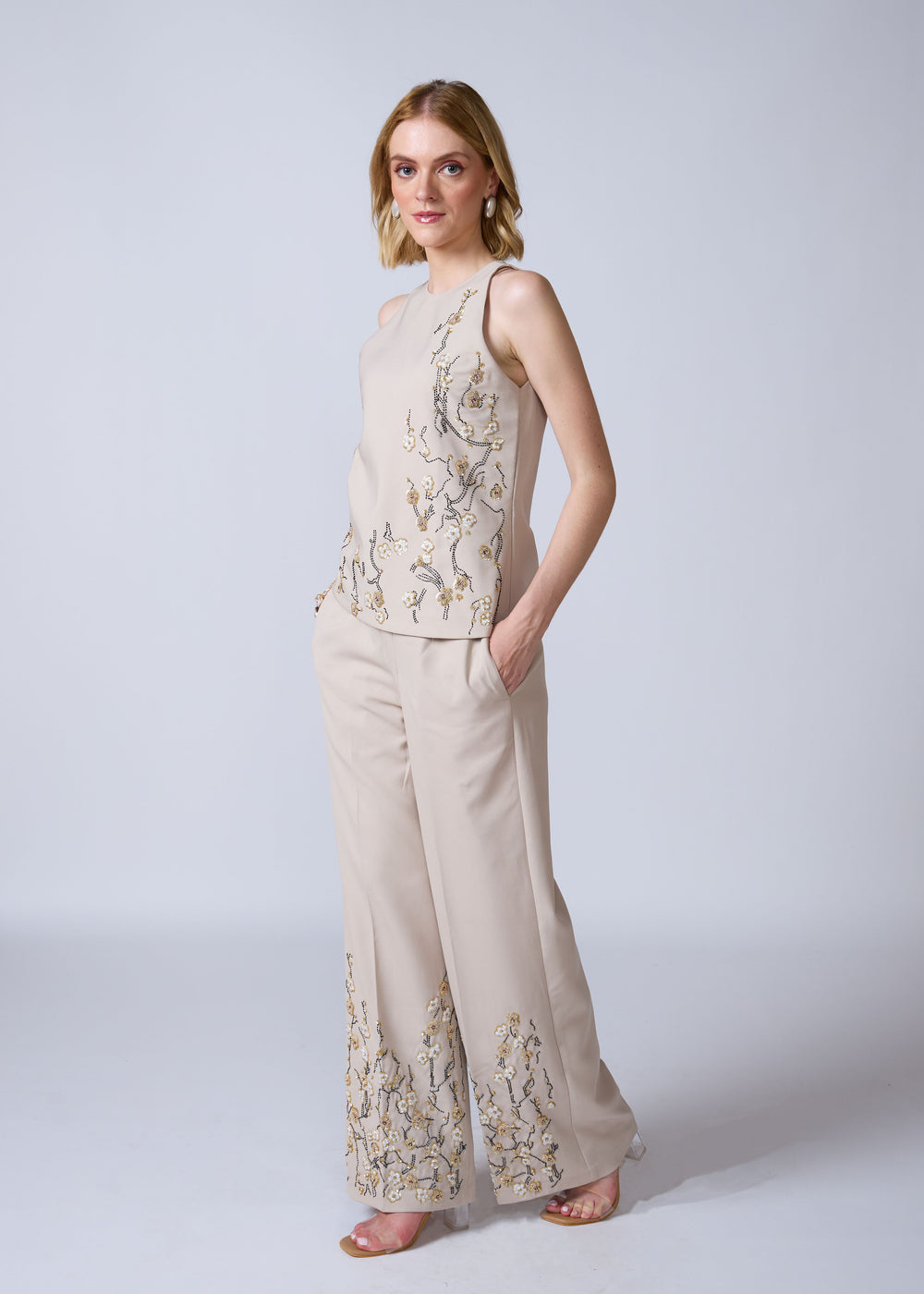 Woman wearing a beige sleeveless top and wide-leg pants with floral embroidery on a white background