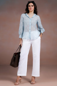 Woman wearing a light blue shirt and white pants, holding a black handbag against a plain background