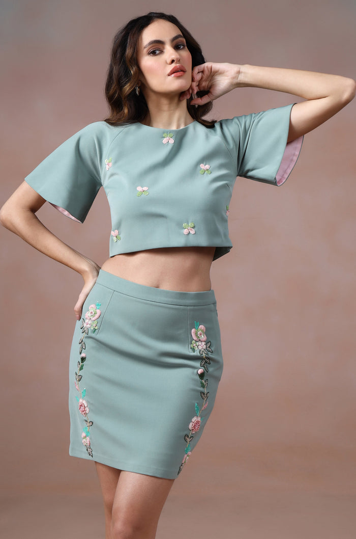 Woman wearing a matching teal top and skirt with floral embroidery against a gradient background