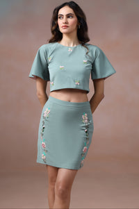 Woman wearing a matching teal top and skirt with floral embroidery against a brown background
