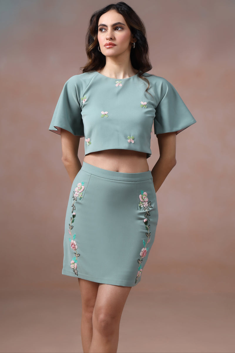 Woman wearing a matching teal top and skirt with floral embroidery against a brown background