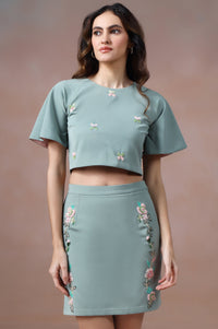 Woman wearing a matching green top and skirt with floral embroidery on a brown background