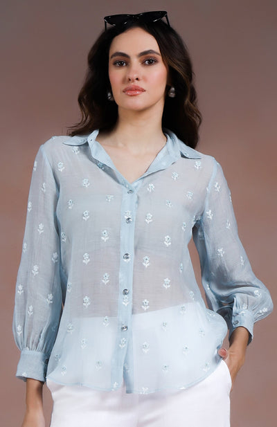 Woman wearing a light blue shirt and white pants, holding a black handbag against a plain background