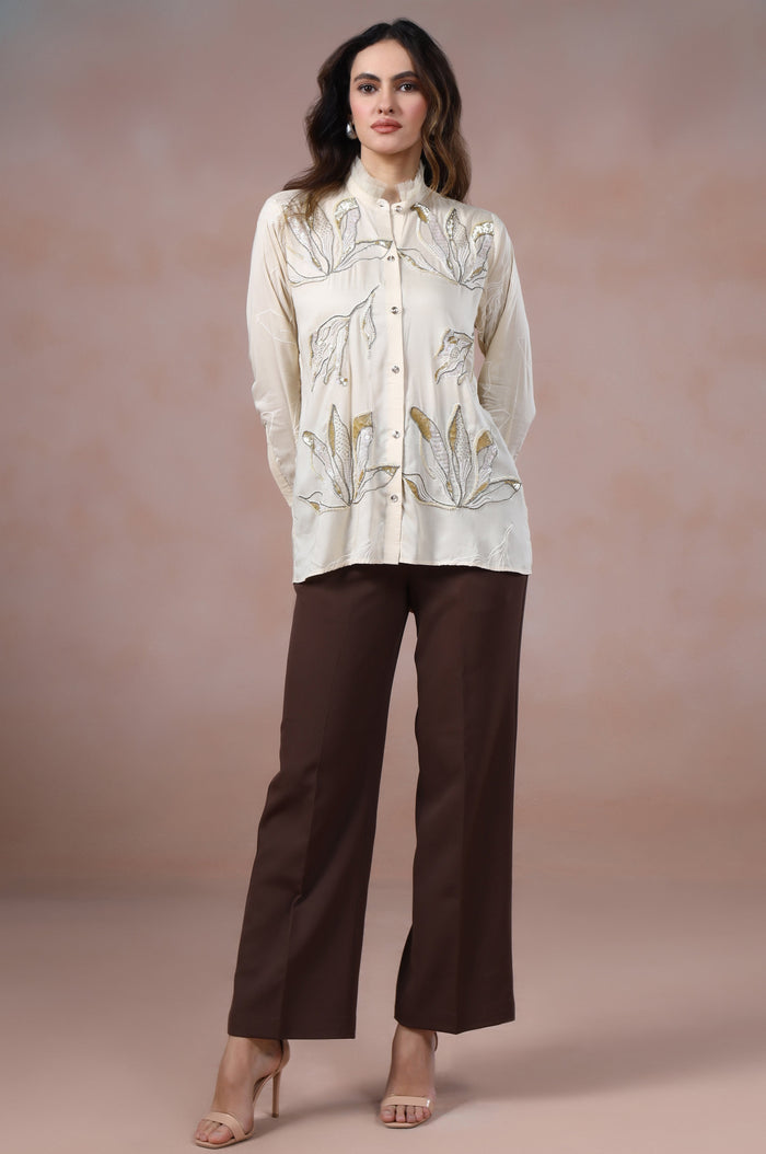 Woman wearing a cream blouse with floral patterns and brown pants against a pink background