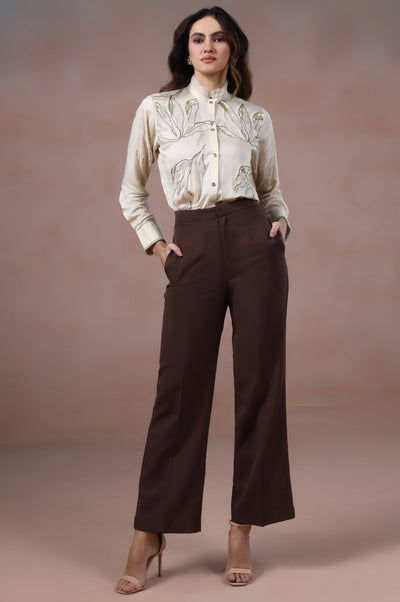 Woman wearing a white blouse and brown pants against a plain background