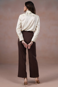 Woman wearing a white blouse and brown pants against a beige background