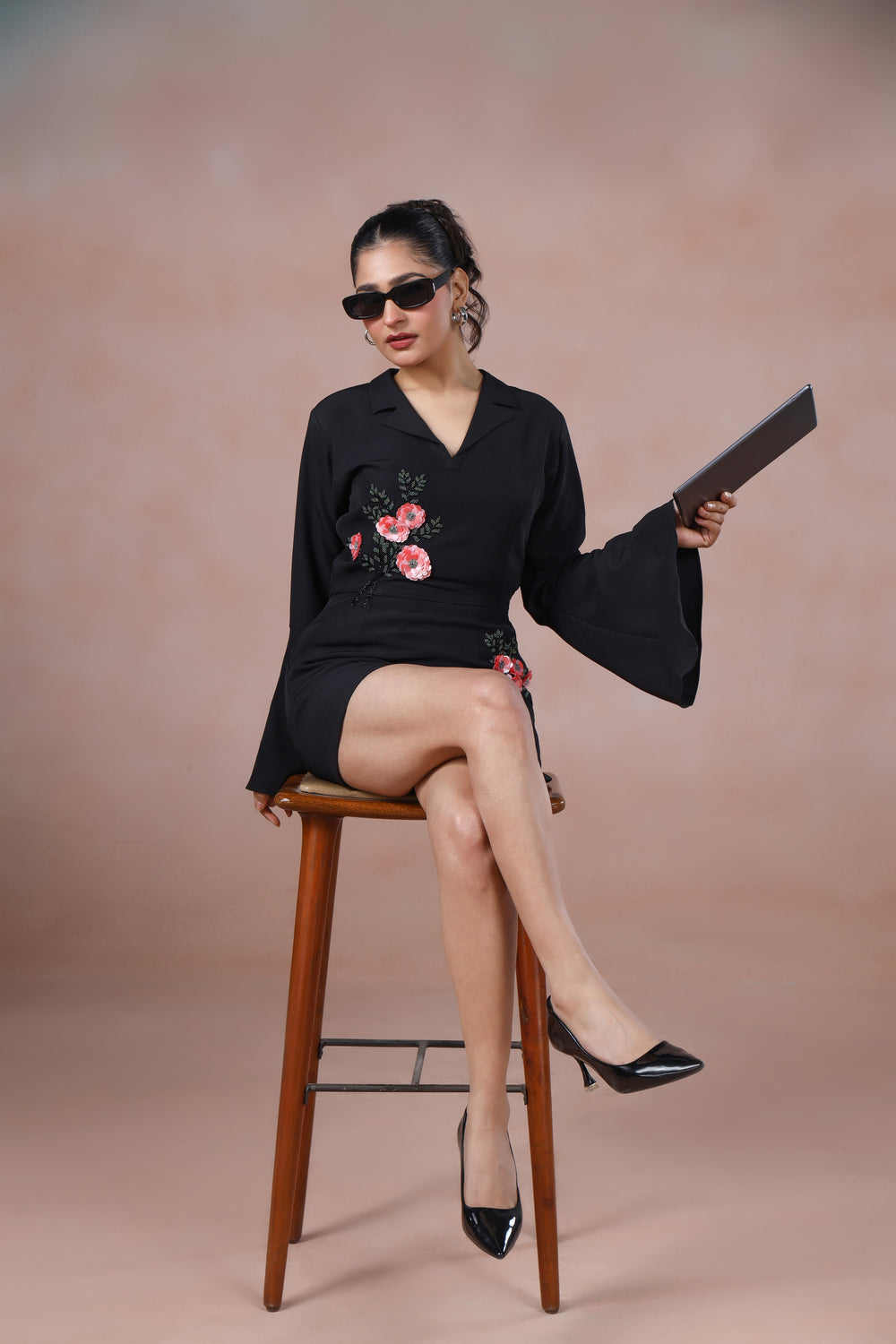 Woman in a black dress with floral embroidery sitting on a wooden stool against a beige background