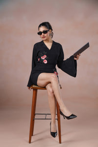 Woman in a black dress with floral embroidery sitting on a wooden stool against a beige background